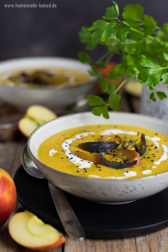 Eine ofengeröstete Kürbissuppe in einer Schüssel, garniert mit gerösteten Apfel- und Zwiebelscheiben, Schwarzkümmel, Sesam und einem Klecks Sahne. Im Hintergrund sind frische Äpfel und grüne Kräuter zu sehen. Die Suppe wird auf einem rustikalen Holztisch serviert.