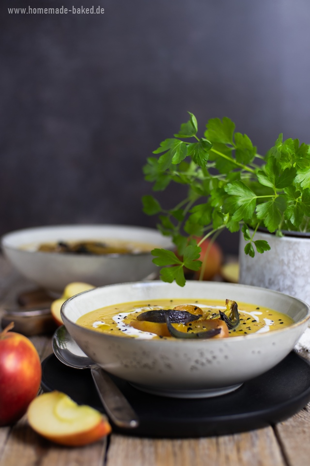 Eine ofengeröstete Kürbissuppe in einer Schüssel, garniert mit gerösteten Apfel- und Zwiebelscheiben, Schwarzkümmel, Sesam und einem Klecks Sahne. Im Hintergrund sind frische Äpfel und grüne Kräuter zu sehen. Die Suppe wird auf einem rustikalen Holztisch serviert.