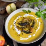 Eine ofengeröstete Kürbissuppe in einer Schüssel, garniert mit gerösteten Apfel- und Zwiebelscheiben, Schwarzkümmel, Sesam und einem Klecks Sahne. Im Hintergrund sind frische Äpfel und grüne Kräuter zu sehen. Die Suppe wird auf einem rustikalen Holztisch serviert.