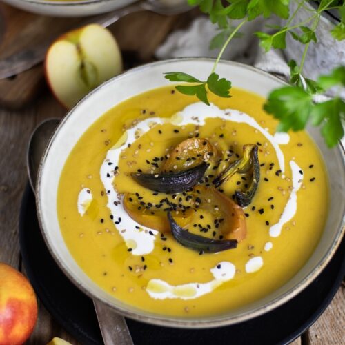 Eine ofengeröstete Kürbissuppe in einer Schüssel, garniert mit gerösteten Apfel- und Zwiebelscheiben, Schwarzkümmel, Sesam und einem Klecks Sahne. Im Hintergrund sind frische Äpfel und grüne Kräuter zu sehen. Die Suppe wird auf einem rustikalen Holztisch serviert.