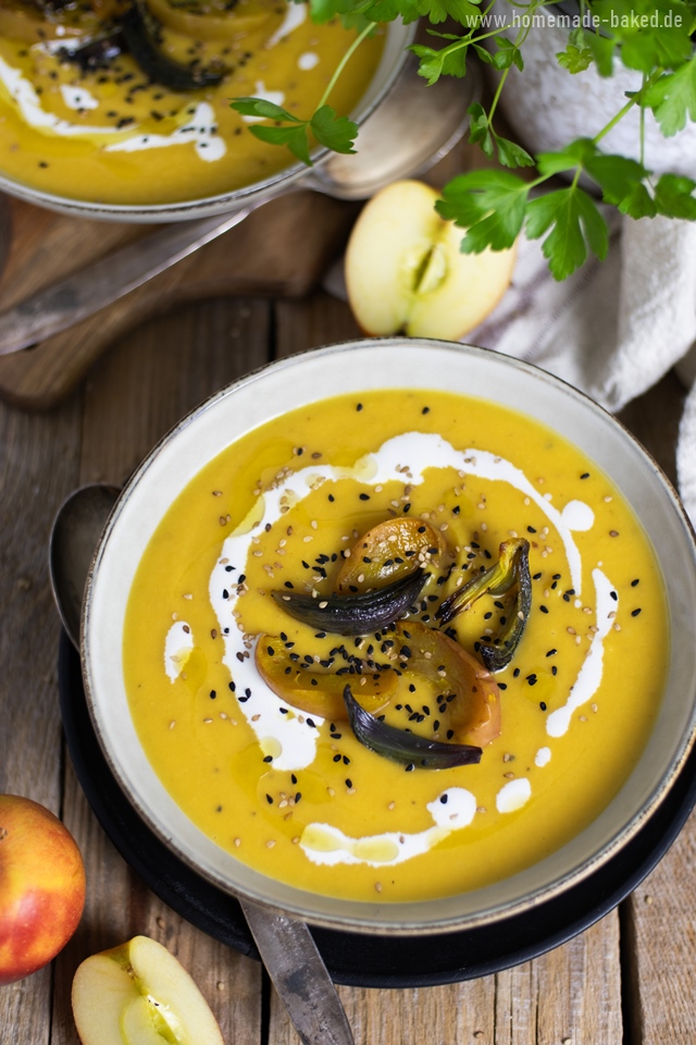 Eine ofengeröstete Kürbissuppe in einer Schüssel, garniert mit gerösteten Apfel- und Zwiebelscheiben, Schwarzkümmel, Sesam und einem Klecks Sahne. Im Hintergrund sind frische Äpfel und grüne Kräuter zu sehen. Die Suppe wird auf einem rustikalen Holztisch serviert.
