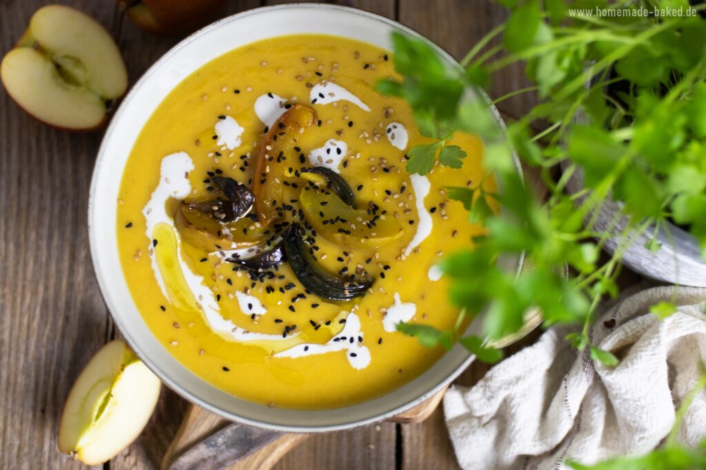 Eine ofengeröstete Kürbissuppe in einer Schüssel, garniert mit gerösteten Apfel- und Zwiebelscheiben, Schwarzkümmel, Sesam und einem Klecks Sahne. Im Hintergrund sind frische Äpfel und grüne Kräuter zu sehen. Die Suppe wird auf einem rustikalen Holztisch serviert.