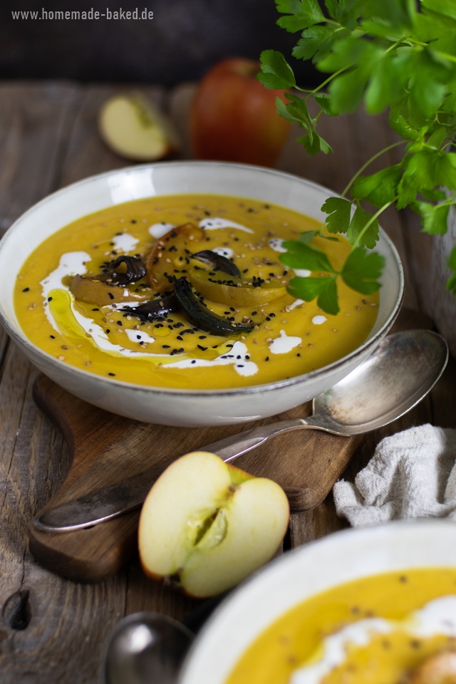 Eine ofengeröstete Kürbissuppe in einer Schüssel, garniert mit gerösteten Apfel- und Zwiebelscheiben, Schwarzkümmel, Sesam und einem Klecks Sahne. Im Hintergrund sind frische Äpfel und grüne Kräuter zu sehen. Die Suppe wird auf einem rustikalen Holztisch serviert.
