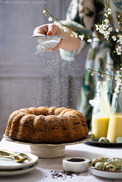 Saftiger Eierlikör-Gugelhupf mit Schokostreuseln auf einer Kuchenplatte, wird gerade mit Puderzucker bestäubt