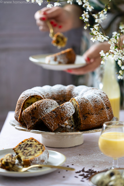 Aufgeschnittener, saftiger Eierlikör-Gugelhupf mit Schokostreuseln auf einer Kuchenplatte, bestäubt mit Puderzucker - im Hintergrund wird ein Stück davon gegessen