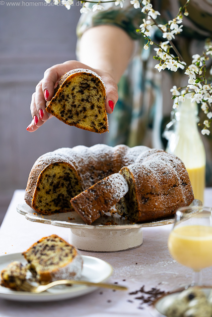Aufgeschnittener, saftiger Eierlikör-Gugelhupf mit Schokostreuseln auf einer Kuchenplatte, bestäubt mit Puderzucker - ein Stück wird hochgenommen