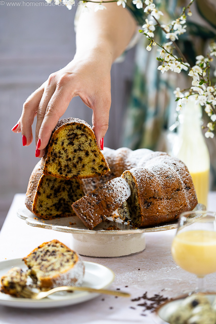 Aufgeschnittener, saftiger Eierlikör-Gugelhupf mit Schokostreuseln auf einer Kuchenplatte, bestäubt mit Puderzucker - ein Stück wird hochgenommen