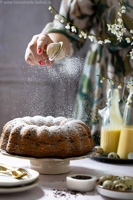 Saftiger Eierlikör-Gugelhupf mit Schokostreuseln auf einer Kuchenplatte, wird gerade mit Puderzucker bestäubt
