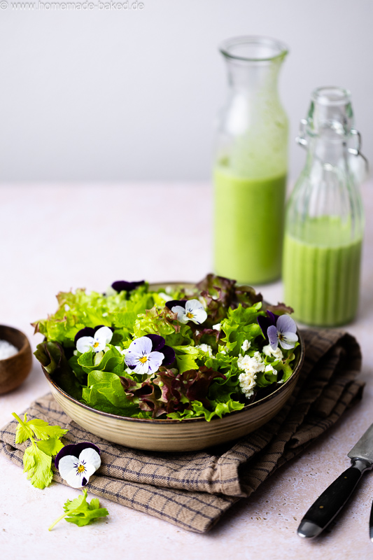 Frischer Salat mit Bärlauch-Buttermilch-Dressing, Feta und essbaren Blumen. Im Hintergrund stehen zwei Flaschen mit Dressing.