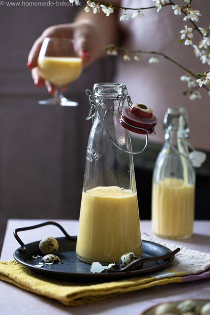 Selbstgemachter Eierlikör mit ganzen Eiern aus dem Thermomix in zwei Flaschen, im Hintergrund wird ein Glas mit Eierlikör in der Hand gehalten.