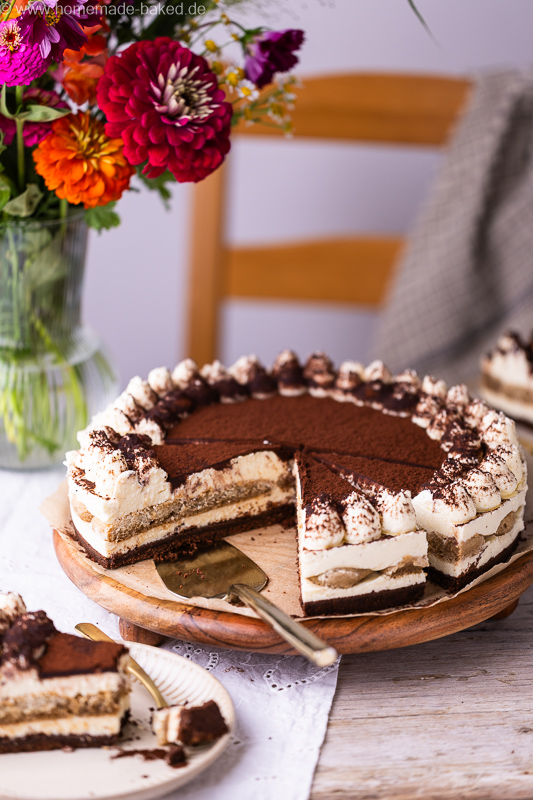 Angeschnittener No Bake Tiramisu Käsekuchen mit cremiger Füllung und feiner Kakaoschicht, im Hintergrund ein Strauß bunter Blumen