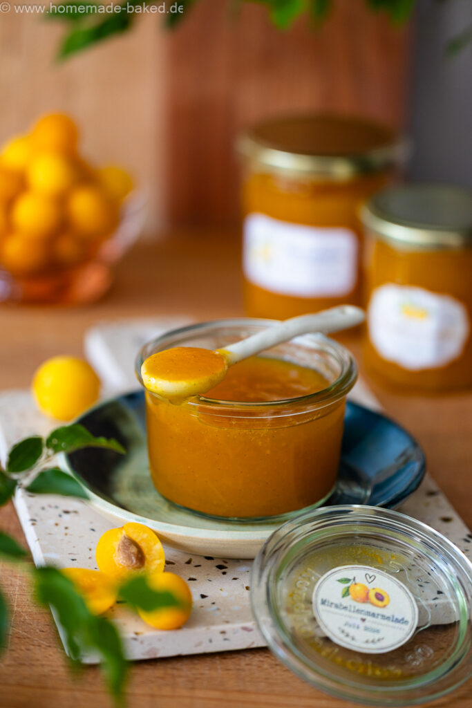 Mirabellenmarmelade mit Vanille im geöffneten Glas, mit einem Löffel voll fruchtiger Marmelade, dahinter zwei geschlossene Marmeladengläser.