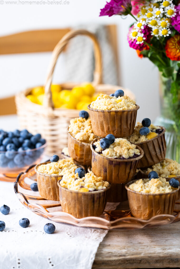 thermomix streuselmuffins zu einer pyramide gestapelt und mit heidelbeeren dekoriert. Auf einem tisch mit frischen blumen, im hintergrund ein obstkorb
