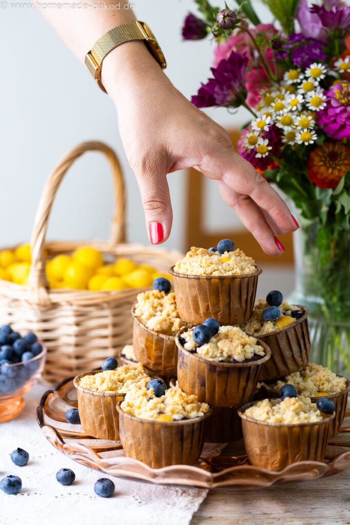 thermomix streuselmuffins zu einer pyramide gestapelt und mit heidelbeeren dekoriert. Auf einem tisch mit frischen blumen, im hintergrund ein obstkorb. Nach dem oberen muffin wird gegriffen.
