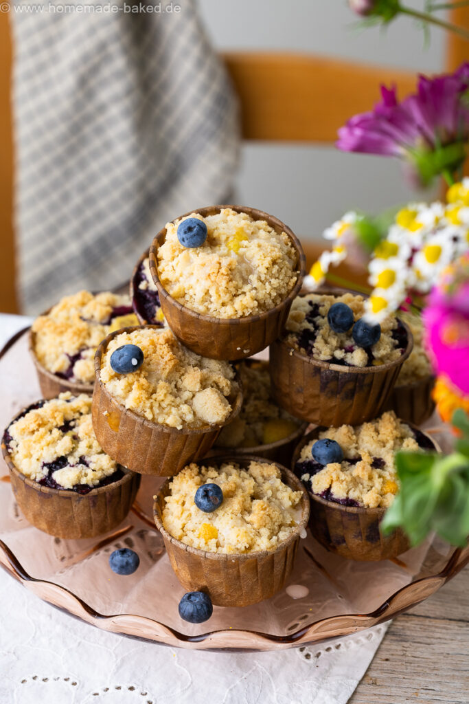 thermomix streuselmuffins zu einer pyramide gestapelt und mit heidelbeeren dekoriert. Auf einem tisch mit frischen blumen