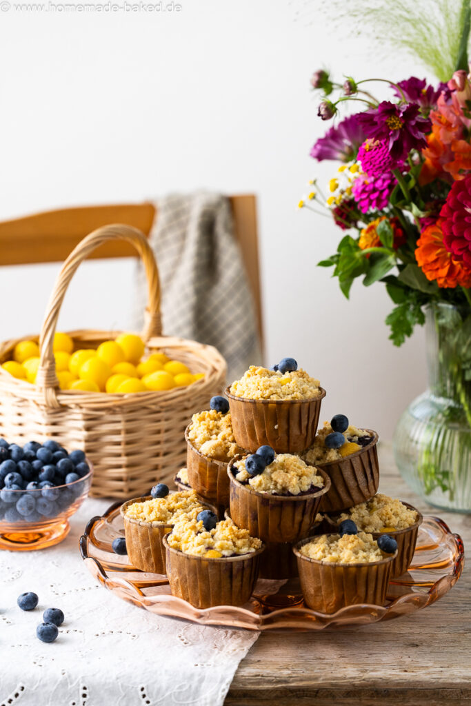 thermomix streuselmuffins zu einer pyramide gestapelt und mit heidelbeeren dekoriert. Auf einem tisch mit frischen blumen, im hintergrund ein obstkorb