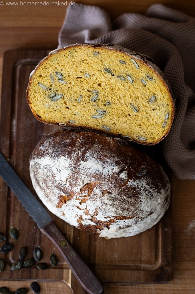 Kürbis-Sauerteigbrot mit Kürbiskerne, in der Mitte aufgeschnitten und auf ein Brett gelegt, so dass die saftige Krume und die Kruste gleichzeitig zu sehen sind.