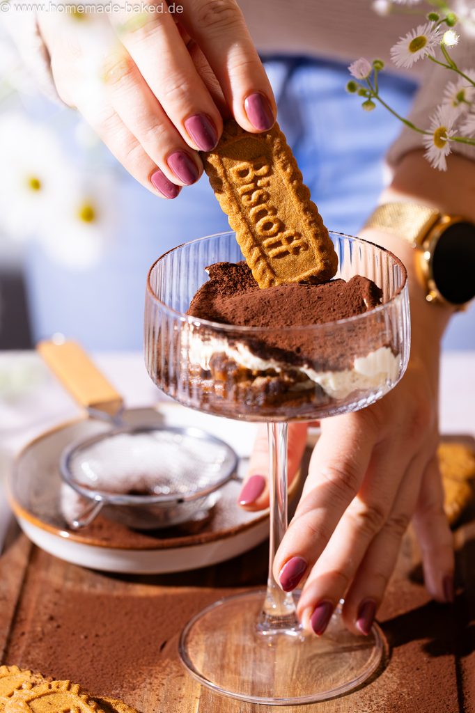 Lotus Biscoff Tiramisu im Glas, in einem hohen Champagnerkelch serviert, eine Hand am Glas-Stiel. Die andere Hand steckt einen ganzen Keks hinein.